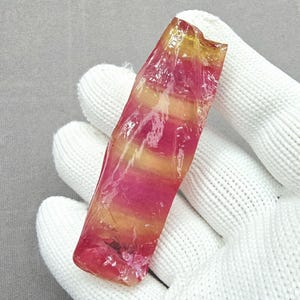 May include: A raw, elongated gemstone with a vibrant color gradient. The stone transitions from a deep pink at the base to a golden yellow at the top. The crystal is held in a white glove, showcasing its natural, unpolished texture.