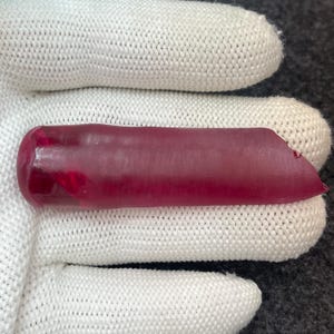 May include: A translucent, elongated, deep red gemstone with a rounded end and a chipped edge. The gemstone is held in a white knitted glove, with a dark gray background.