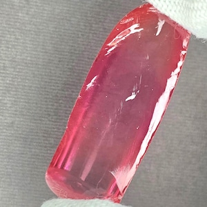 May include: A rectangular, translucent, pink gemstone with rounded edges. The gemstone has a polished surface with visible internal inclusions and a slight color gradient. The gem is held by a white fabric.