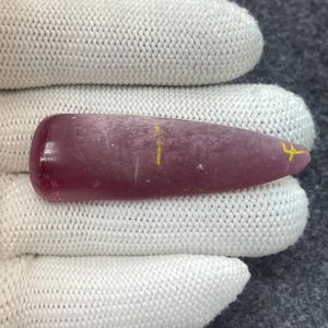 Peut inclure: Une pierre précieuse polie, allongée, rouge-violacé. La pierre a une extrémité arrondie et se termine en pointe. Des marques jaunes sont visibles sur la surface. Le bijou est tenu par un gant blanc tricoté.