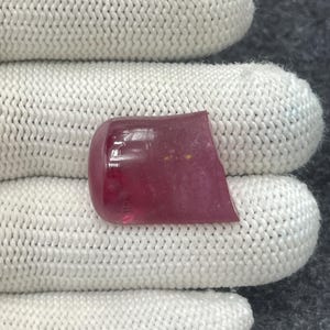 May include: A polished, cabochon-cut gemstone with a deep red hue. The stone has a rounded, smooth top and a flat, irregular bottom. The gemstone is held against a white, knitted fabric.