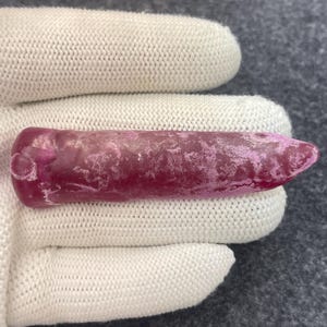 May include: A raw, elongated gemstone with a deep magenta hue. The stone has a textured surface with lighter pinkish-white patches. The shape is cylindrical with a pointed end, held in a white glove.