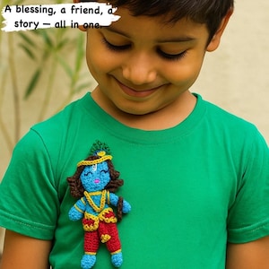 May include: A crocheted Krishna doll in blue, red, and yellow is pinned to a green t-shirt. The doll has a serene expression and is adorned with a crown and jewelry. Text above reads: "A blessing, a friend, a story - all in one."