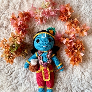 Könnte beinhalten: Handgefertigte Häkelpuppe einer Gottheit, mit blauem Gesicht und Körper, roter Hose und gelbem Schärpe. Die Puppe hat schwarzes Haar, eine gelbe Krone und ist mit einem Blumengirlande geschmückt. Sie hält einen kleinen braunen Topf. Orange und rosa Blumen umgeben die Puppe.