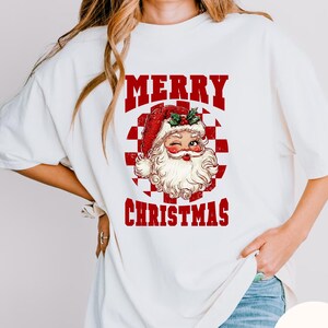 May include: White t-shirt featuring a vintage Santa Claus graphic with the words "MERRY CHRISTMAS" in red block letters. Santa is winking and wearing a red hat. The background is a red and white checkerboard pattern.