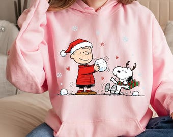 Könnte beinhalten: Hellrosa Kapuzenpullover mit einer Cartoon-Illustration von Charlie Brown und Snoopy im Schnee. Charlie Brown trägt einen Weihnachtsmannhut und einen roten Mantel, Snoopy hat ein Geweih und einen Schal. Schneeflocken und Herzen sind verstreut.