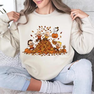 May include: Cream-colored sweatshirt featuring a fall-themed graphic. The design includes cartoon characters playing in a pile of autumn leaves, surrounded by pumpkins and falling leaves. The graphic is in shades of brown, orange, and yellow.