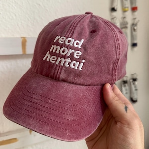 Könnte beinhalten: Burgunderfarbene Baseballkappe mit den Worten "read more hentai" in Weiß gestickt. Die Kappe hat einen gebogenen Schirm und einen Used-Look.