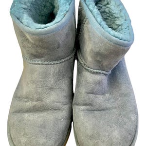 May include: A pair of ankle-height boots featuring a light gray suede exterior and a soft, light blue faux fur lining. The boots have a rounded toe, visible stitching along the seams, and a thick, flat sole, providing a simple and functional design.