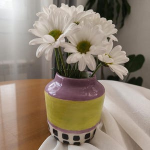 Puede incluir: Un jarrón de cerámica con borde morado, una banda central ancha de color verde lima y una base con un patrón de cuadrícula en blanco y negro. El jarrón está lleno de un ramo de margaritas blancas con centros amarillos, mostrando un diseño vibrante.