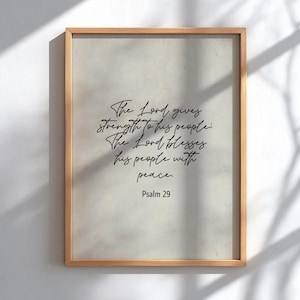 Op de afbeelding: Een ingelijste print met een lichtbeige achtergrond en een houten frame. De print bevat een elegant zwart schrift dat luidt: "De Heer geeft kracht aan zijn volk: De Heer zegent zijn volk met vrede. Psalm 29."