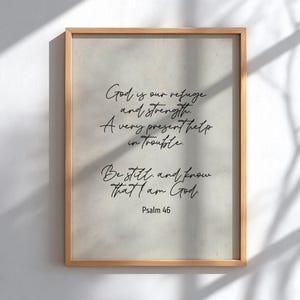 May include: A framed print with a light beige background and a wooden frame. The print features elegant black script with the text "God is our refuge and strength. A very present help in trouble. Be still, and know that I am God. Psalm 46."