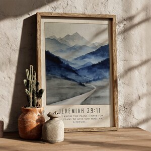 Könnte beinhalten: Ein gerahmter Aquarelldruck mit einer Berglandschaft in Blau- und Grautönen. Der Druck enthält den Text "JEREMIAH 29:11" und ein Zitat über Hoffnung und Zukunft. Ein Kaktus in einem Terrakottatopf und eine kleine Vase befinden sich im Vordergrund.