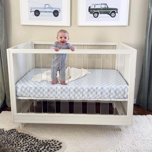 Peut inclure: Un lit bébé blanc avec des côtés en acrylique transparent et un drap-housse à carreaux bleu et blanc. Un bébé en barboteuse grise se tient debout dans le lit, souriant. Deux impressions encadrées de camions vintage sont accrochées au mur.