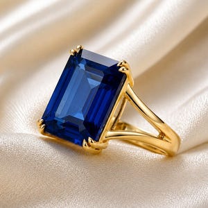 May include: A gold ring featuring a large, rectangular, emerald-cut deep blue gemstone held by four prongs. The band is crafted from polished yellow gold and splits into two distinct arms as it connects to the setting of the central stone.