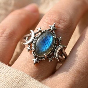 May include: A silver ring featuring an oval-shaped iridescent blue labradorite gemstone at the centre. The setting is decorated with two crescent moons on the sides and small four-pointed star accents surrounding the central stone.