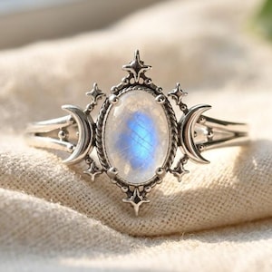 Op de afbeelding: Een zilveren ring met een ovale maansteen met een blauwe iriserende glans. De zetting is versierd met halve maanmotieven aan beide zijden en kleine vierpuntige steraccenten boven en onder de centrale edelsteen.