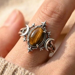 May include: A silver ring featuring an oval-shaped tiger's eye gemstone at the centre. The band is decorated with two crescent moon motifs on the sides and small four-pointed star accents surrounding the central stone setting.