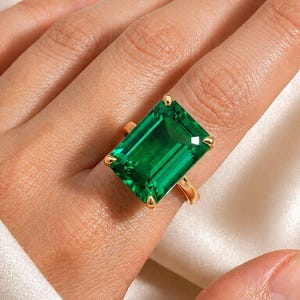 May include: A gold ring featuring a large, rectangular, emerald-cut green gemstone held in place by four prongs. The vibrant green stone is set on a simple, polished gold band that encircles the finger, showcasing a clear, faceted surface.