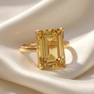 May include: A gold ring with a large, rectangular, yellow gemstone. The ring's band and setting are gold, creating a cohesive design. The gemstone has a faceted cut, enhancing its brilliance. The ring is set against a white background.