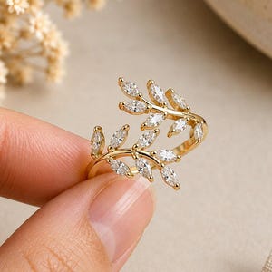 May include: A gold-toned open ring featuring two overlapping branch-like designs. Each branch is adorned with multiple marquise-cut clear stones arranged to resemble leaves. The metal band is polished and slender, creating a delicate botanical aesthetic.