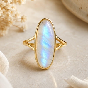 May include: A gold ring with an oval moonstone. The moonstone displays iridescent colours of white, blue and yellow. The ring has a split shank design.