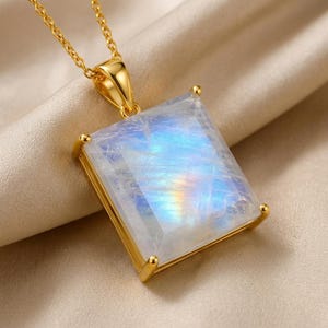 May include: A square-cut moonstone pendant set in a polished gold-tone metal frame, suspended from a matching gold-tone chain. The gemstone displays a translucent white appearance with iridescent blue and rainbow flashes across its faceted surface.