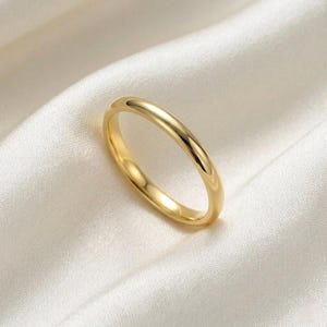 Pode incluir: Um anel de ouro simples e polido. O anel tem uma banda lisa e arredondada e uma superfície brilhante e refletora. É apresentado sobre um fundo branco e sedoso, realçando a sua cor dourada e o seu design minimalista.