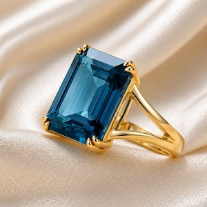 May include: A gold ring featuring a large, rectangular, emerald-cut deep blue gemstone held by four prongs. The band has a split-shank design that tapers toward the setting, showcasing a polished metallic finish and a vibrant, saturated blue stone.