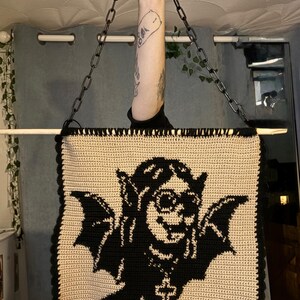 May include: A crocheted tote bag in black and cream. The bag displays a black graphic of a winged creature with a cross pendant. It is suspended by a black chain and a wooden dowel. The bag is handmade.