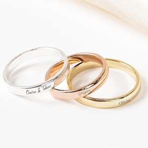 May include: Three personalised rings in silver, rose gold, and gold. The silver ring is engraved with "Claire & Tobias", the rose gold ring with "Gregory", and the gold ring with "Olivia". The rings are displayed on a white surface.