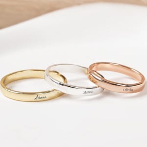 Peut inclure: Trois bagues personnalisées en or, argent et or rose. Chaque bague est gravée d'un nom : Ariana, Marcus et Olivia. Les bagues sont présentées sur une surface blanche.