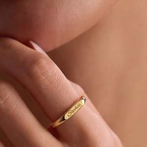 Puede incluir: Un anillo de banda de oro pulido con una superficie lisa y redondeada que presenta el nombre Sophia grabado en una delicada fuente cursiva. El anillo tiene un diseño minimalista con un grosor constante y un acabado metálico brillante.