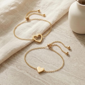 Puede incluir: Dos pulseras doradas con dijes de corazón y cadenas ajustables. Una tiene un corazón calado, la otra un corazón macizo. Descansan sobre una superficie clara, con un pequeño jarrón y flores secas en el fondo.