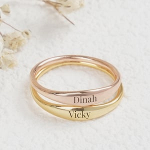 May include: Stack of two personalized rings, one rose gold and one gold, with the names "Dinah" and "Vicky" engraved on them. The rings are on a white textured surface, with delicate white flowers in the background.