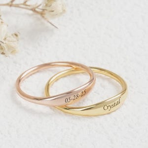 May include: Two delicate rings, one rose gold and one gold, are intertwined on a textured white surface. The rose gold ring is engraved with "05-28-25", while the gold ring is inscribed with "Crystal". The rings are simple bands, perfect for everyday wear.