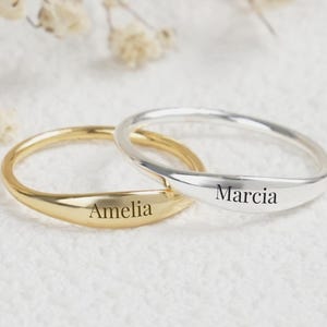 May include: Two personalized rings, one gold and one silver, each engraved with a name. The gold ring says "Amelia," and the silver ring says "Marcia." The rings are simple bands with a slight curve.