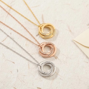 Peut inclure: Trois colliers personnalisés en or, or rose et argent. Chaque collier comporte trois anneaux imbriqués sur une chaîne délicate. Les anneaux sont gravés de noms et de dates. Le fond est une surface texturée blanc cassé.