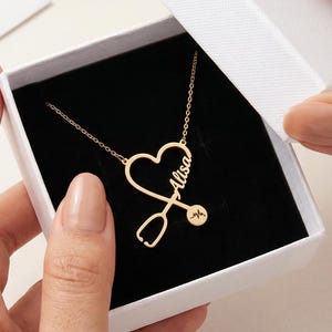 Könnte beinhalten: Eine goldfarbene Halskette mit herzförmigem Anhänger und Stethoskop-Design. Der Name "Alisa" ist auf dem Herzen eingraviert. Die Halskette wird in einer weißen Schachtel mit schwarzem Innenraum präsentiert.