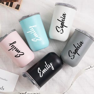 Op de afbeelding: Een assortiment van vijf ge&iuml;soleerde bekers in pastelkleuren: roze, lichtblauw, wit en grijs, plus een zwarte. Elke beker heeft een zilveren rand en een naam in cursief schrift. De namen zijn "Emily" en "Sophia".