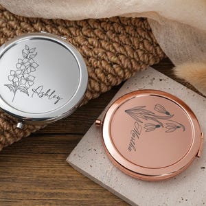 May include: Two round compact mirrors. One is silver with a floral design and the name "Ashley" in script. The other is rose gold with a floral design and the name "Martha" in script. Both mirrors are open.