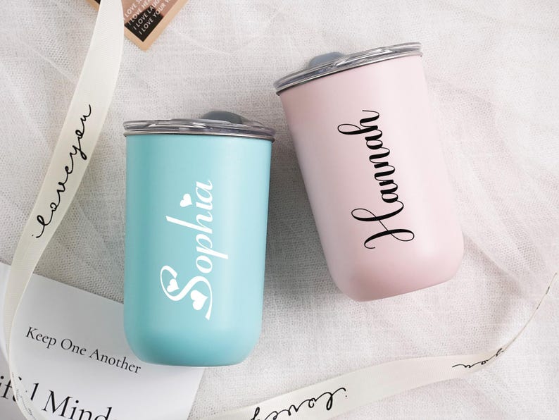 Op de afbeelding: Twee gepersonaliseerde bekers, &eacute;&eacute;n lichtblauw en &eacute;&eacute;n roze, elk met een naam in elegant schrift. De blauwe beker zegt "Sophia" met hartaccenten, terwijl de roze "Hannah" zegt. Beide hebben zilveren deksels.