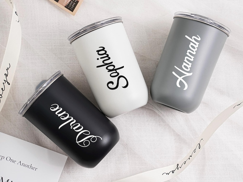 Op de afbeelding: Drie gepersonaliseerde bekers in zwart, wit en grijs. Elke beker heeft een zilveren rand en een naam in een sierlijk lettertype: Darlene, Sophia en Hannah. De bekers staan op een wit oppervlak met een lint met de tekst "I love you."