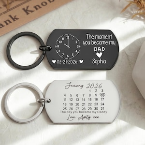 Peut inclure: Deux porte-clés personnalisés. L'un, noir, présente un design d'horloge avec le texte "The moment you became my DAD" et une date. L'autre, argenté, a un design de calendrier et le texte "The day you became my Daddy".