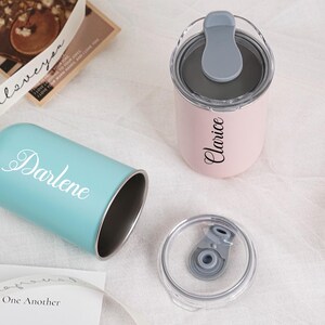 Op de afbeelding: Twee gepersonaliseerde bekers, &eacute;&eacute;n lichtblauw met "Darlene" in witte letters en een roze met "Clarice". Beide hebben zilveren interieurs en grijze deksels. Een doorzichtig deksel is ook zichtbaar.