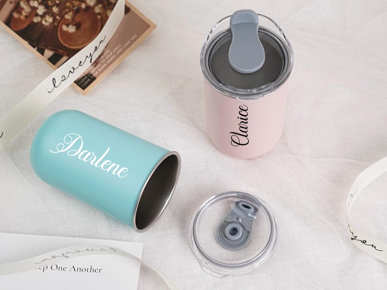 Op de afbeelding: Twee gepersonaliseerde bekers, &eacute;&eacute;n lichtblauw met "Darlene" in witte letters en een roze met "Clarice". Beide hebben zilveren interieurs en grijze deksels. Een doorzichtig deksel is ook zichtbaar.