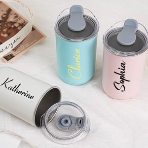 Op de afbeelding: Drie gepersonaliseerde roestvrijstalen bekers in pastelkleuren: lichtblauw, roze en wit. Elke beker heeft een naam erop: "Clarice", "Sophia" en "Katherine". De bekers hebben grijze deksels met een klapdeksel.
