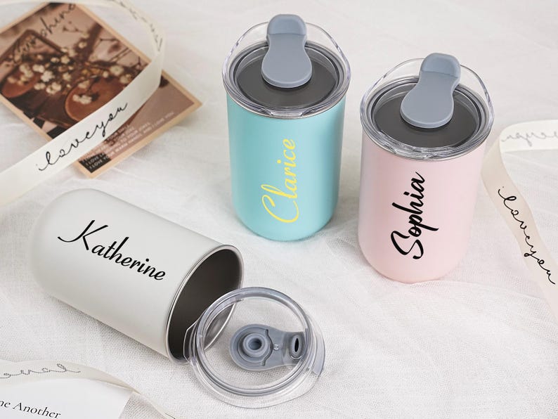 Op de afbeelding: Drie gepersonaliseerde roestvrijstalen bekers in pastelkleuren: lichtblauw, roze en wit. Elke beker heeft een naam erop: "Clarice", "Sophia" en "Katherine". De bekers hebben grijze deksels met een klapdeksel.