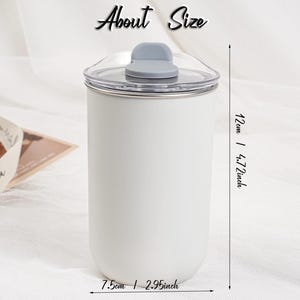 Op de afbeelding: Een witte beker met een doorzichtig deksel en een grijze klapsluiting. De beker is 12 cm hoog en 7,5 cm breed. De tekst "About Size" staat bovenaan de afbeelding.