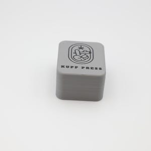Op de afbeelding: Een grijze, rechthoekige pers met de tekst "KUPP PRESS" en een grafisch ontwerp erop. De pers is gemaakt van een stevig materiaal en wordt waarschijnlijk gebruikt voor het persen van kruiden of andere materialen. Het ontwerp bevat een hand die een plant vasthoudt.